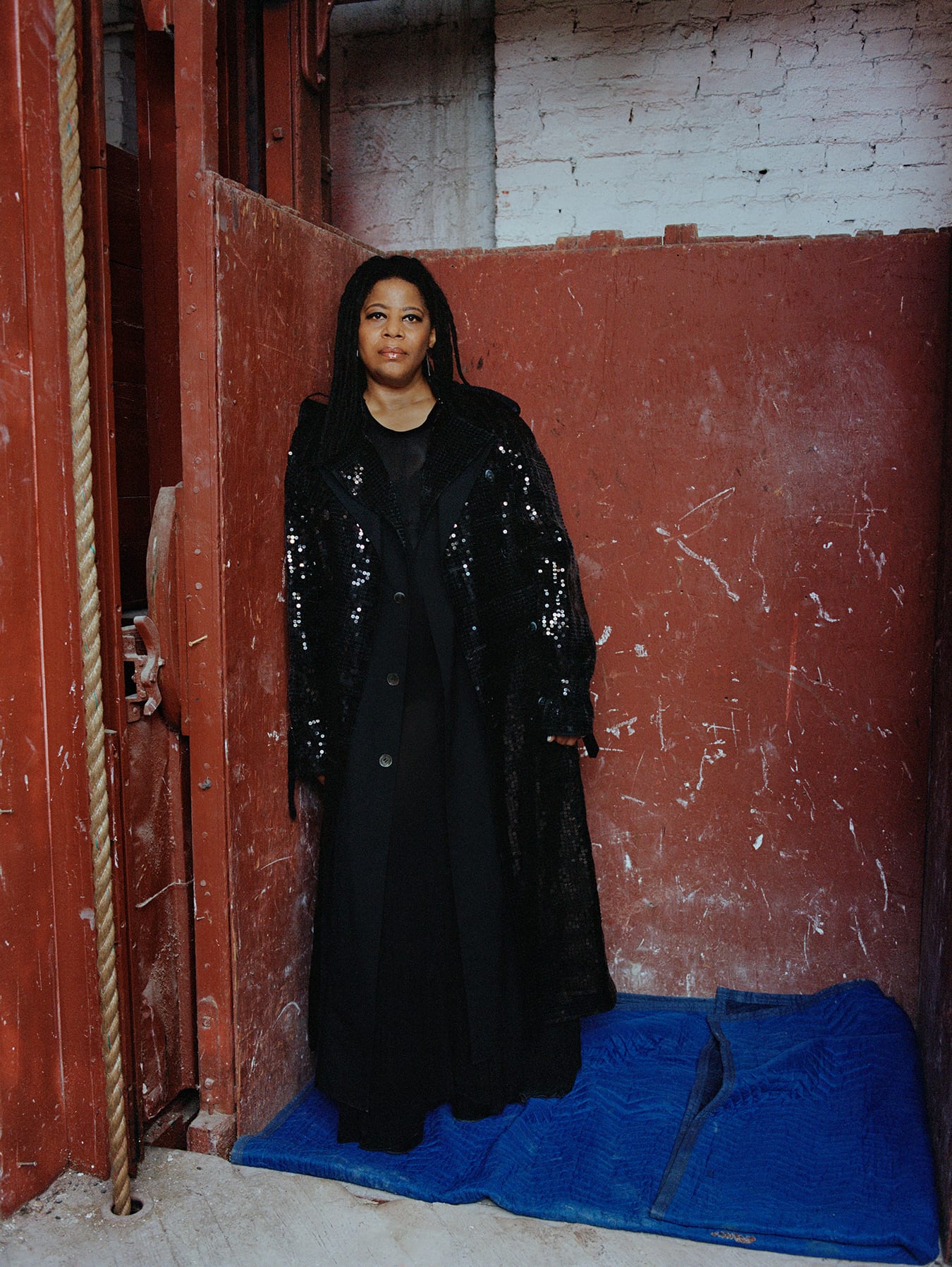 Simone Leigh, a medium-dark-skinned woman with dreadlocks stands in a rust-colored industrial elevator in a sequined black jacket and dress.